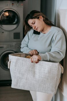 Focused female with bag of clothes having phone conversation while standing in washroom near modern washing machines during housework routine
