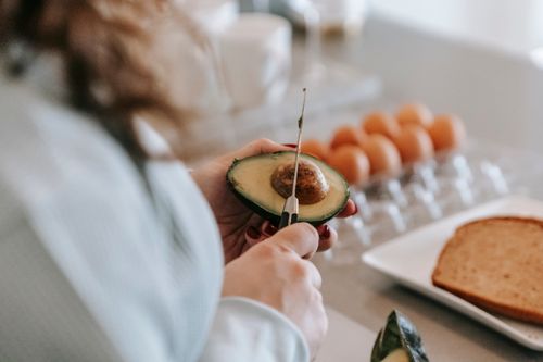 Free Crop anonymous female cook with knife preparing avocado while standing at table with eggs and toasts in light kitchen at home Stock Photo