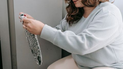 Focused female in casual wear picking dirty clothes from laundry basket while sitting in light room at home during daily housework routine