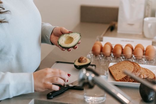 Crop faceless female cook preparing avocado while making toasts at table with sliced sliced bread and eggs in kitchen during breakfast