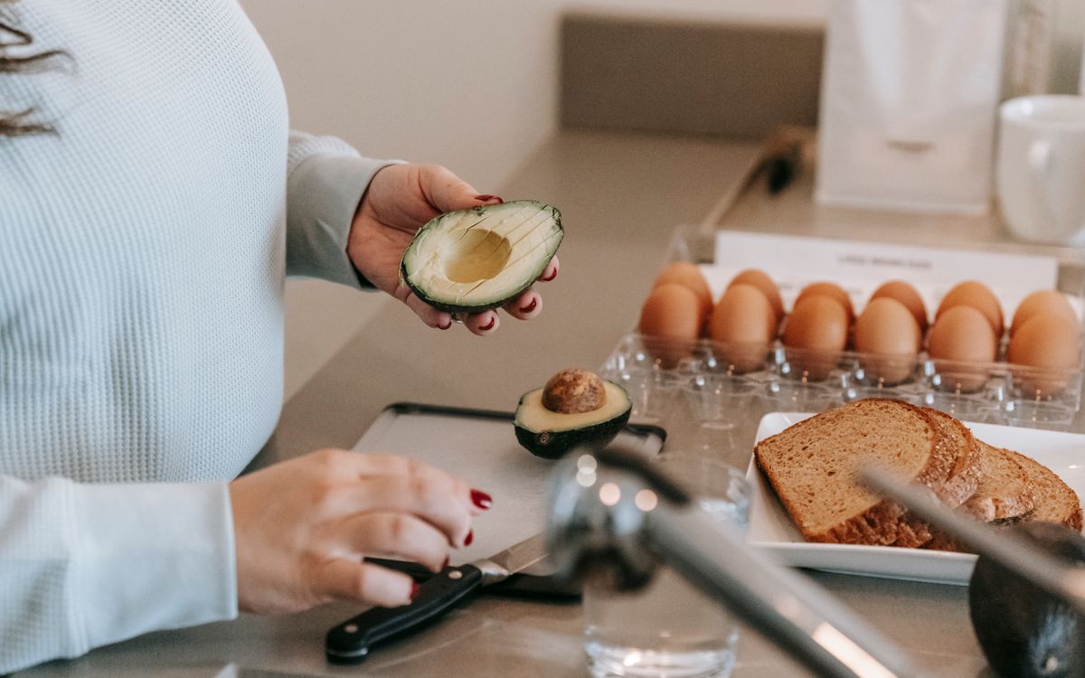 Crop faceless female cook preparing avocado while making toasts at table with sliced sliced bread and eggs in kitchen during breakfast