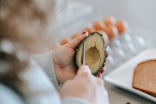 Free Crop anonymous female with knife cutting ripe avocado while preparing toasts at table with eggs and bread on blurred background Stock Photo