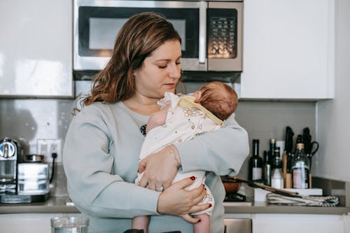 Caring mother embracing cute little infant baby in arms while standing in light modern kitchen with contemporary appliances at home