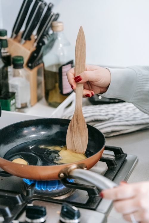 Free Crop anonymous female with wooden spatula stirring frying chicken eggs in pan while cooking on gas stove in light kitchen Stock Photo