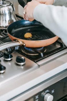 Crop faceless woman breaking raw egg into frying pan on gas stove while preparing breakfast in light kitchen at home