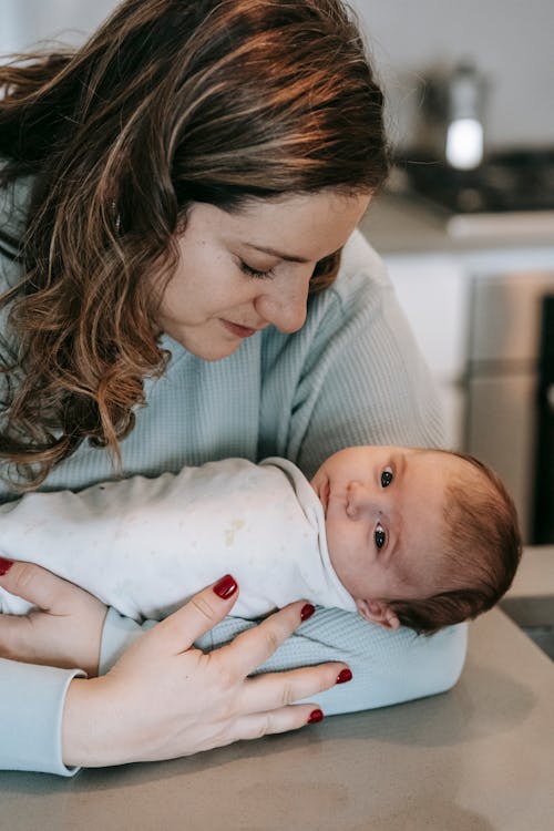 Caring mother with curly hair caressing infant baby on table while standing in light kitchen with modern equipment at home