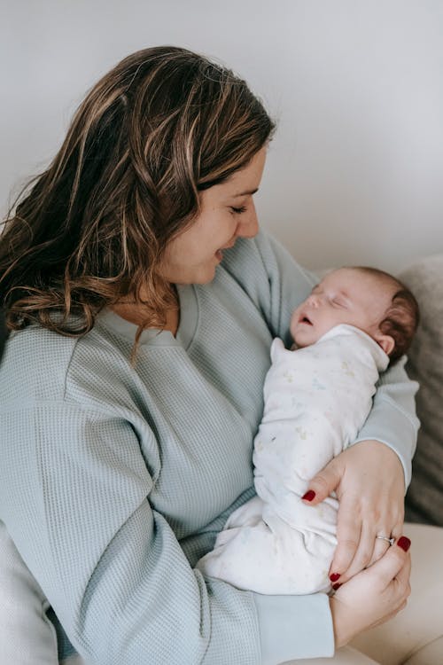 Side view of content female embracing sleeping newborn baby while sitting on sofa near white wall in light living room