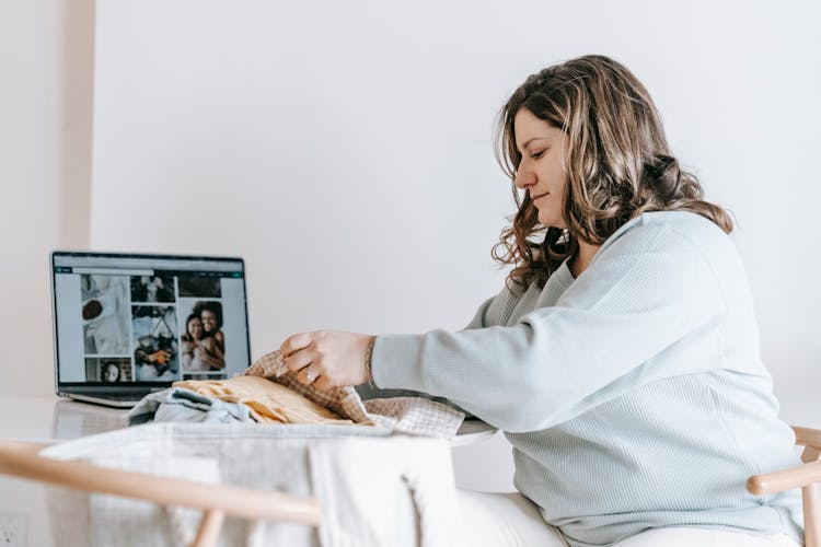 Woman Folding Baby Clothes Near Laptop