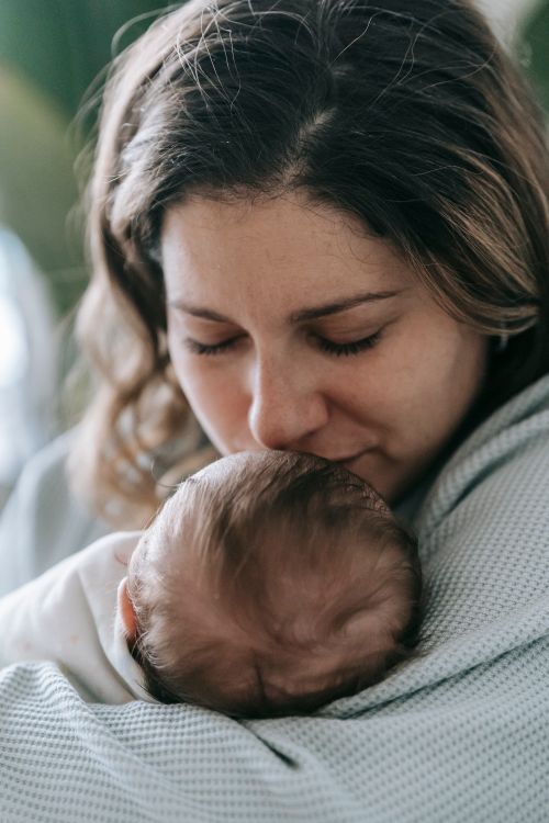 Free Tender mother in casual wear with closed eyes caressing anonymous little infant baby in arms while sitting in light room at home Stock Photo
