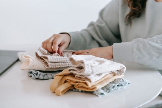 Crop faceless female in casual clothes folded colorful baby garments while sitting at white table in room on white background