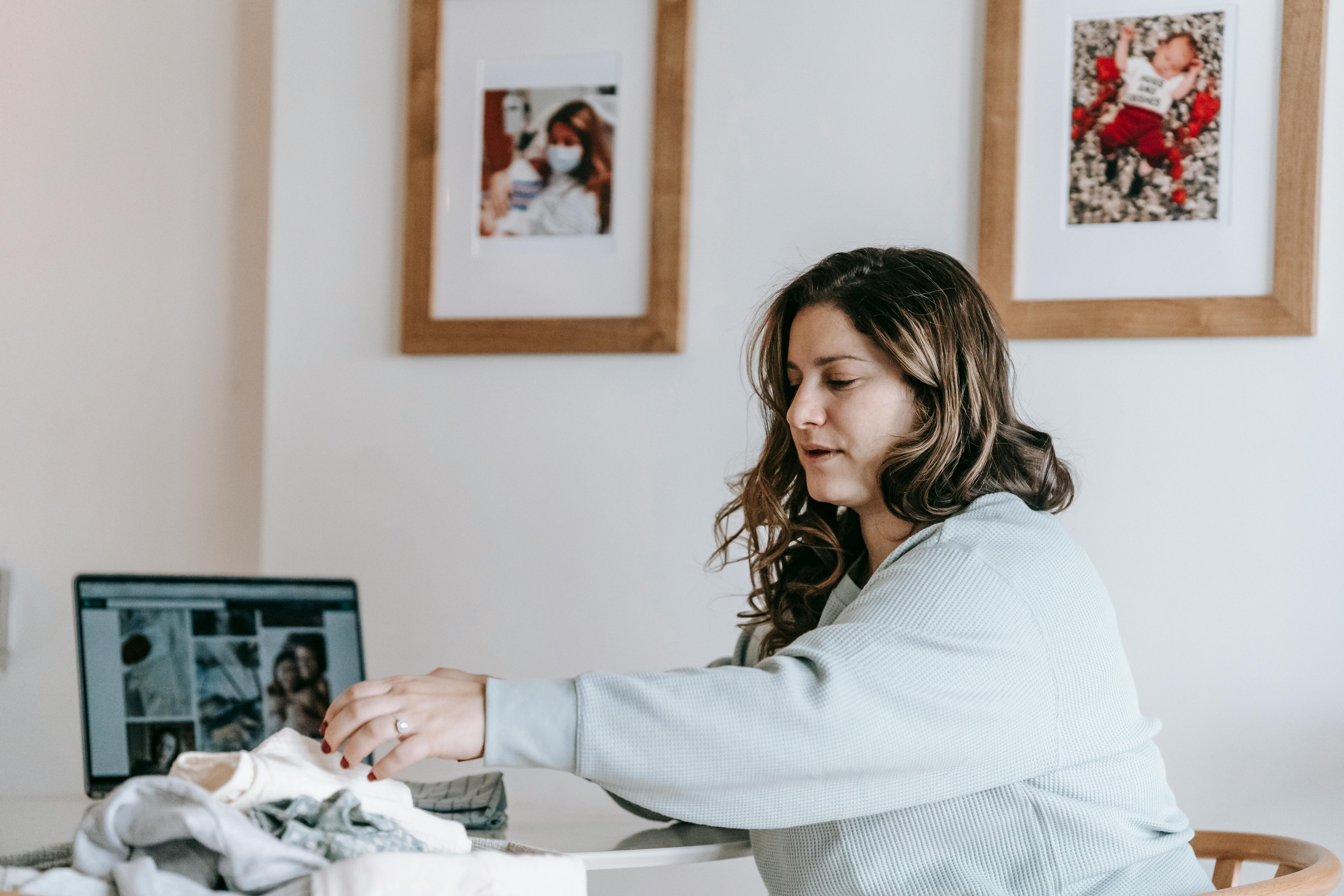Young woman folding clothes at home desk, with laptop open in cozy indoor setting.