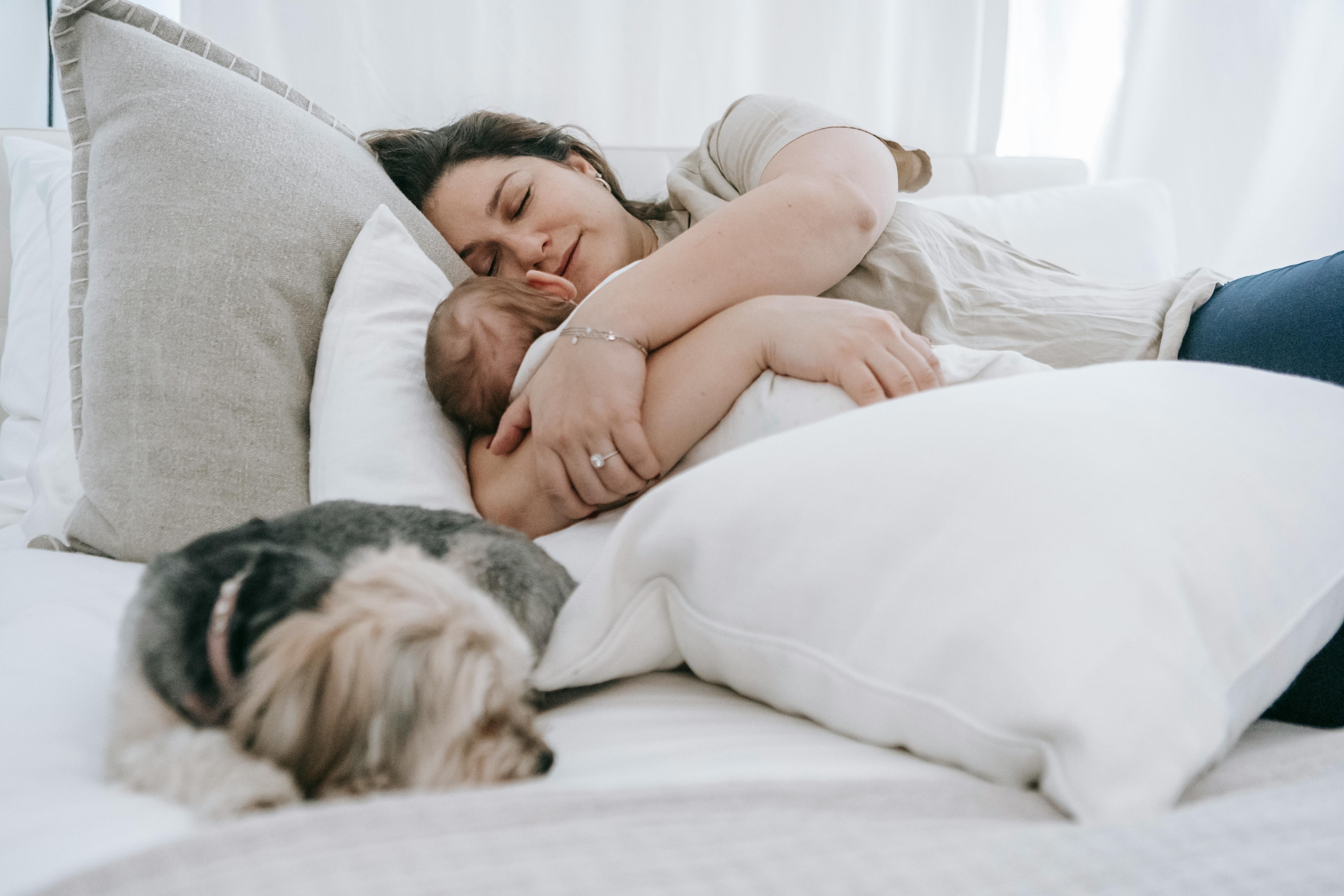 Caring female embracing newborn baby embracing newborn baby while sleeping together on comfortable bed with pillows and morkie dog in bedroom