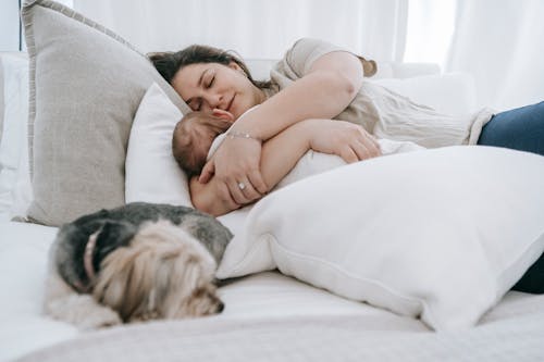 Caring female embracing newborn baby embracing newborn baby while sleeping together on comfortable bed with pillows and morkie dog in bedroom