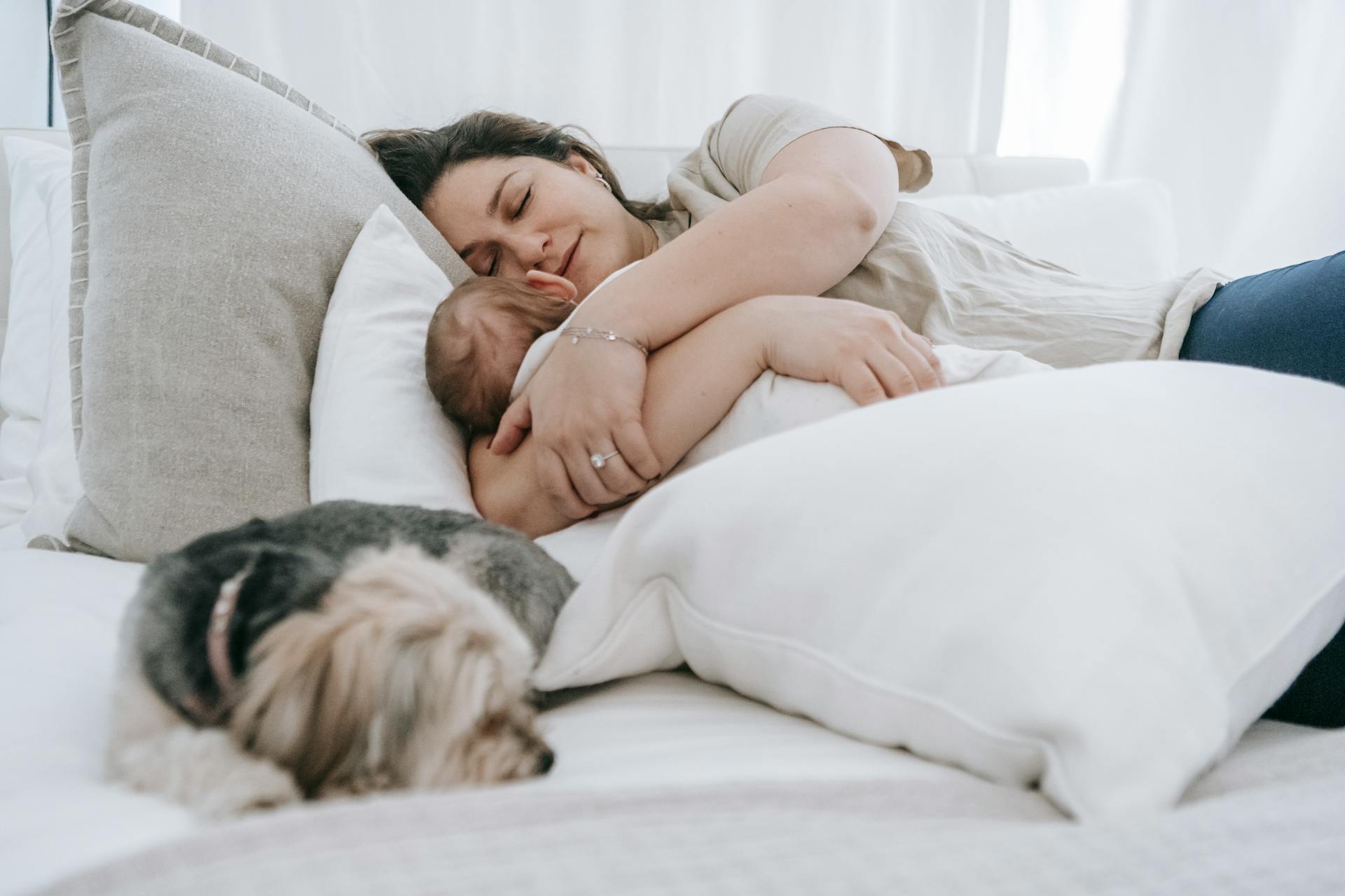 Verzorgende vrouw die een pasgeboren baby omhelst terwijl ze samen slapen op een comfortabel bed met kussens en morkie hond in de slaapkamer