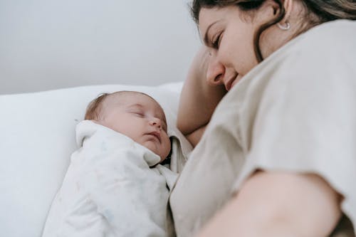 Side view of crop positive caring mother looking at sleeping infant baby while lying on comfortable bed in light bedroom