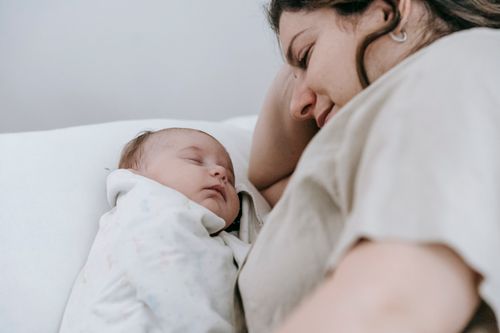 Free Side view of crop positive caring mother looking at sleeping infant baby while lying on comfortable bed in light bedroom Stock Photo