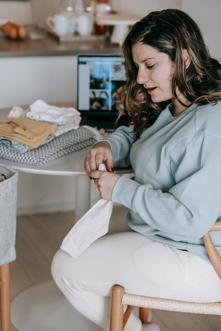 Woman With Folded Baby Clothes Near Laptop On Table