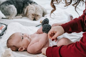 Crop anonymous mother putting on diaper on adorable infant baby wearing anti scratching mittens while lying on comfortable couch near hairy dog in light room
