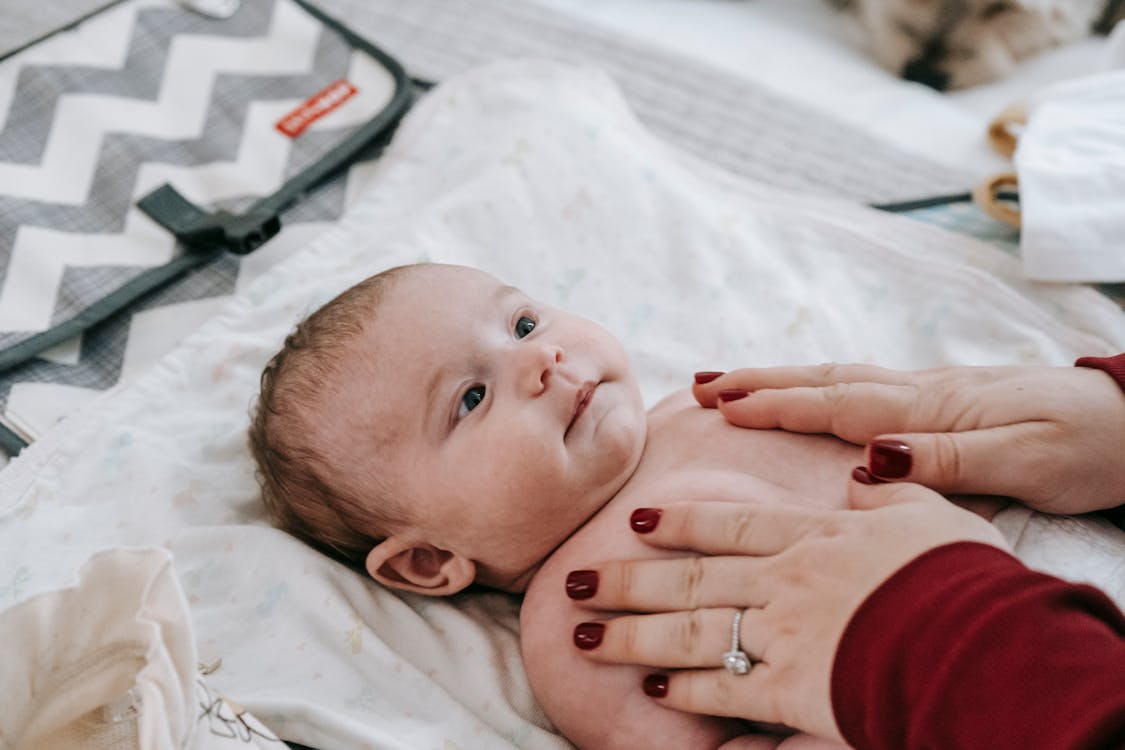 From above of crop anonymous mother with manicure touching adorable infant baby lying on comfortable couch in light room at home