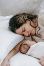 Peaceful mother with closed eyes sleeping with cute little infant baby while lying on comfortable bed in light bedroom at home
