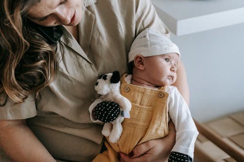 Crop unrecognizable mother in casual wear with cute newborn baby in arms with toy sitting in light room at home