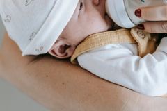 Crop anonymous caring mother feeding newborn hungry baby in arms with bottle in light room on white background at home