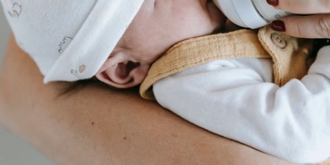 Crop anonymous caring mother feeding newborn hungry baby in arms with bottle in light room on white background at home