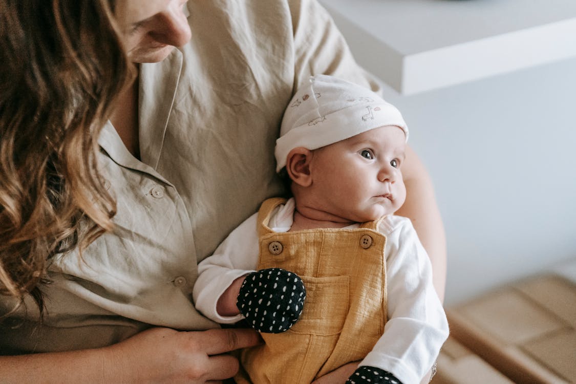 Crop anonymous mother with newborn baby wearing hat and anti scratching mittens in arms sitting in room at home