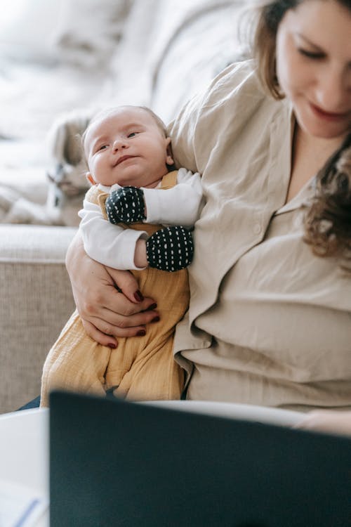 Crop mother with newborn baby in arms surfing internet on netbook while sitting on couch in light room at home