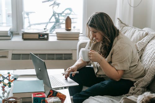 Woman with cup sitting on couch while working on laptop