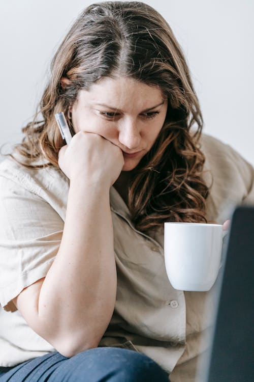 Curve pensive female freelancer leaning on hand sitting near laptop and drinking tea