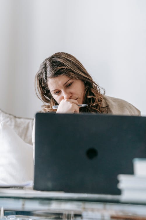 Female worker sitting on sofa at laptop and looking thoughtfully at screen at home