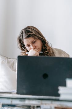 Female worker sitting on sofa at laptop and looking thoughtfully at screen at home