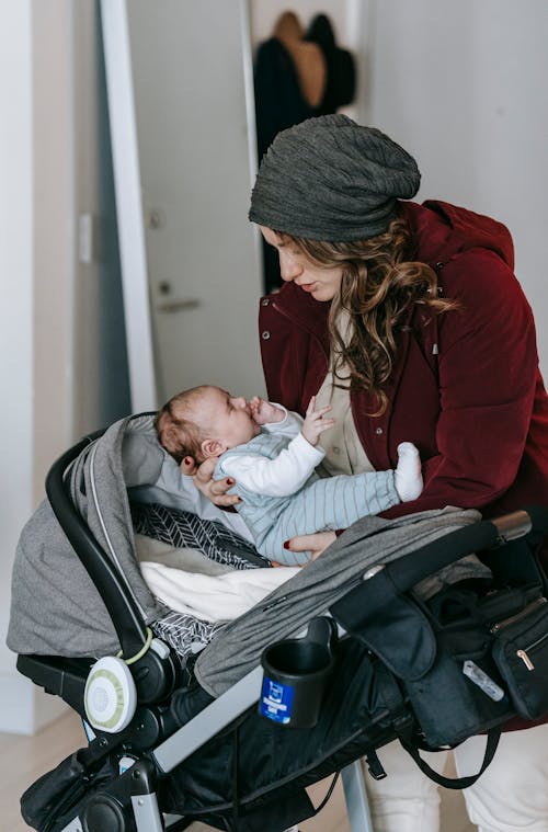 Side view of female in warm clothes taking adorable baby from stroller while standing in room