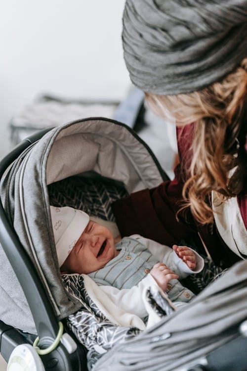 Free Side view of adorable crying baby in casual clothes and hat lying in stroller with closed eyes while crop unrecognizable mother standing near Stock Photo