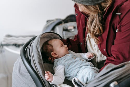 Crop anonymous caring mother in outerwear and hat putting infant baby into gray stroller while standing in light room at home