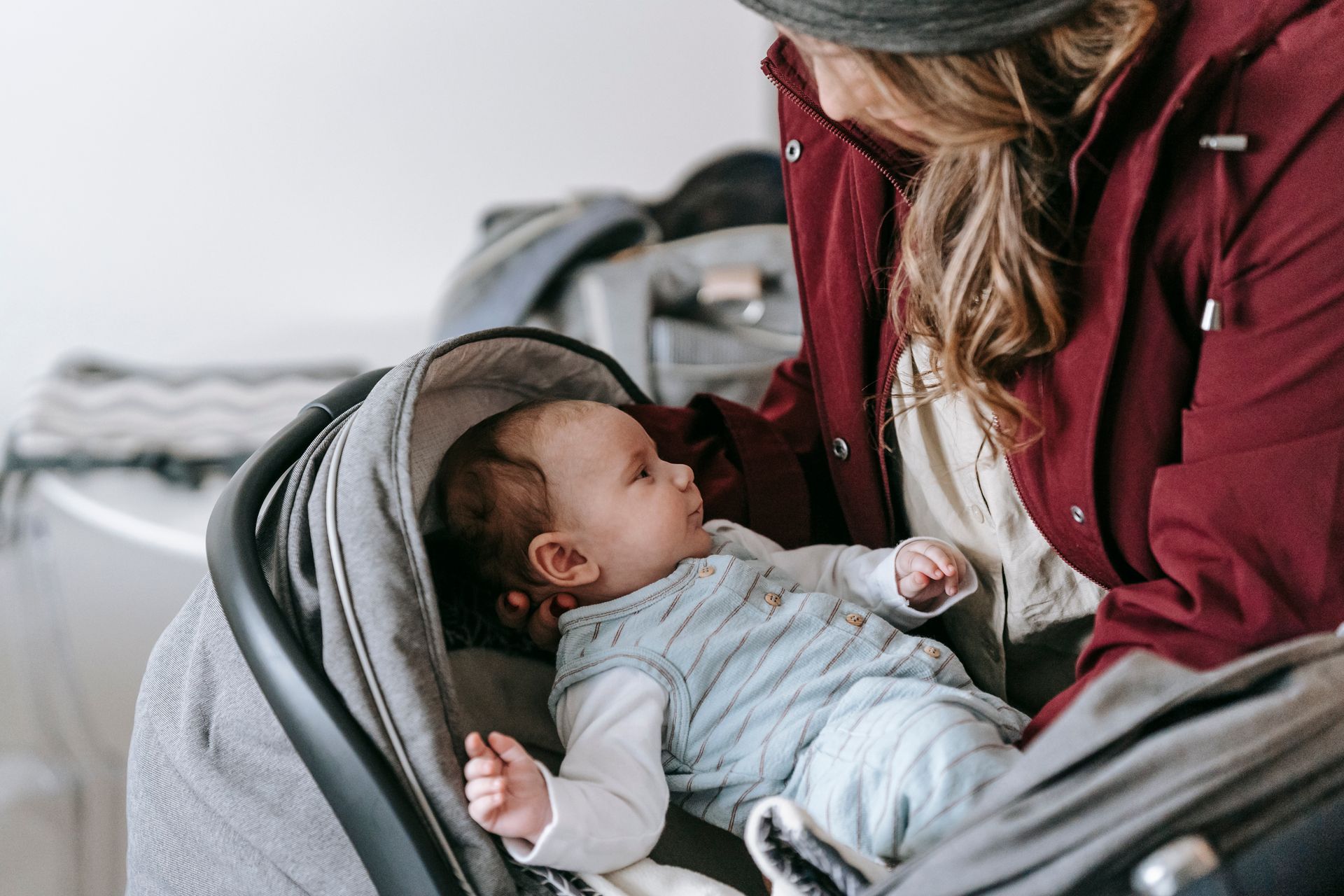 Crop anonymous caring mother in outerwear and hat putting infant baby into gray stroller while standing in light room at home