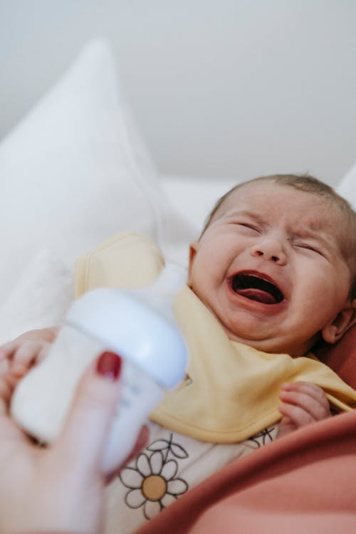 Crop anonymous mother with bottle of milk and screaming upset newborn baby in arms sitting in light room at home