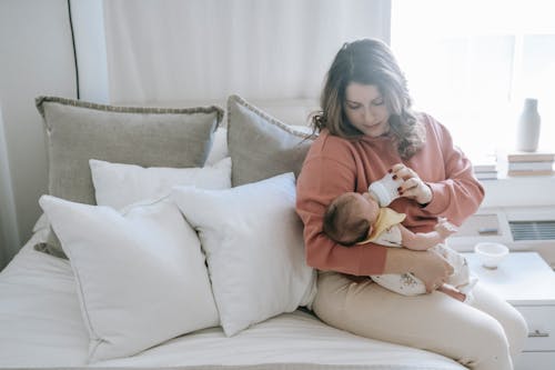 Serious mother feeding infant baby with bottle while sitting on comfortable bed with white pillows in light bedroom with window