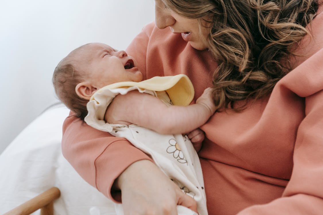 Free Side view of crop anonymous mother consoling crying infant baby in arms while sitting in light room near wall at home Stock Photo