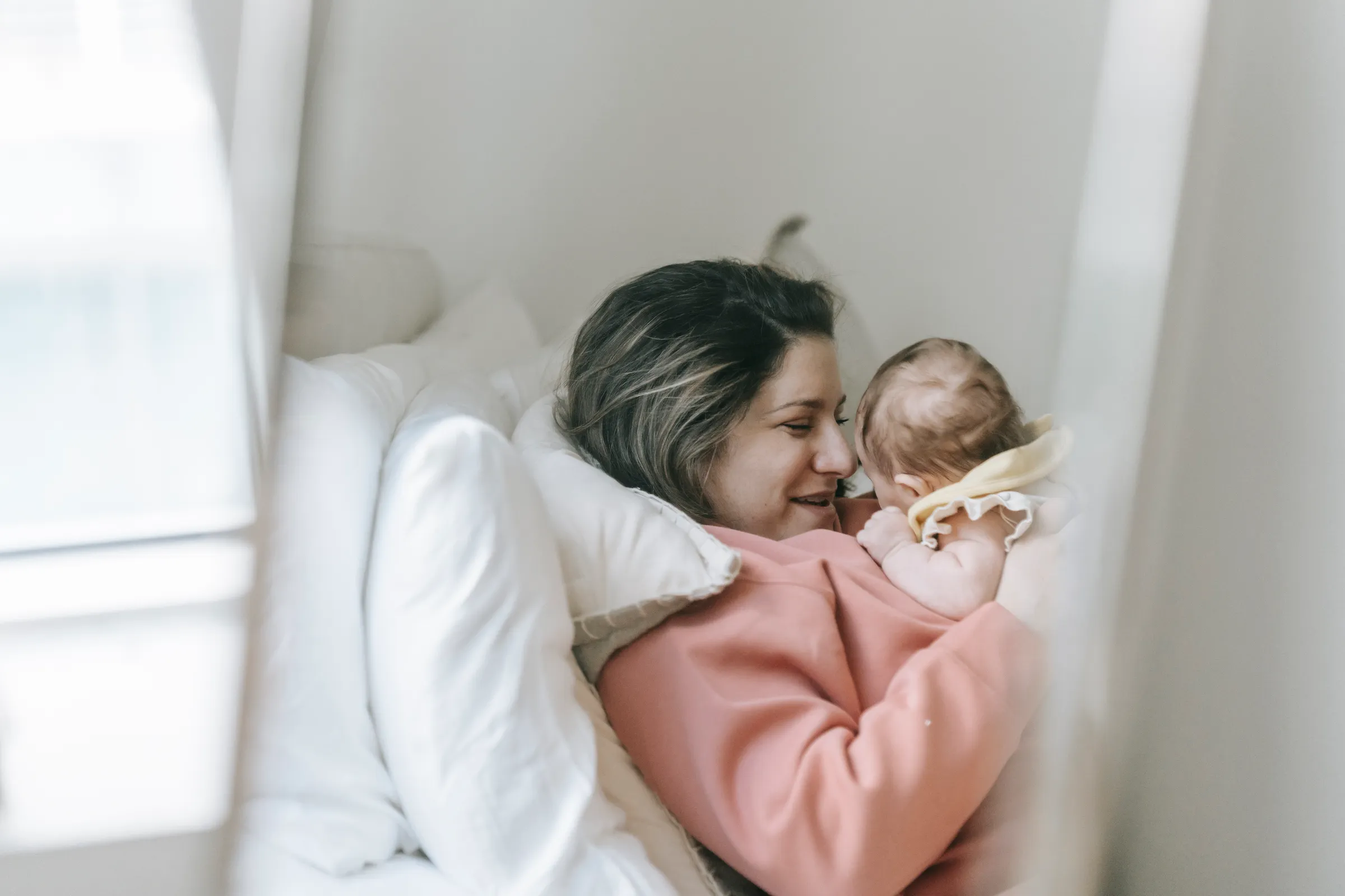 A mother lying face to face with her baby on white bedding, smiling as they look at each other
