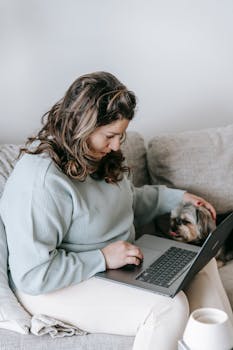 Woman sitting on a couch using a laptop, accompanied by a small dog, showcasing a comfortable work-from-home setting.