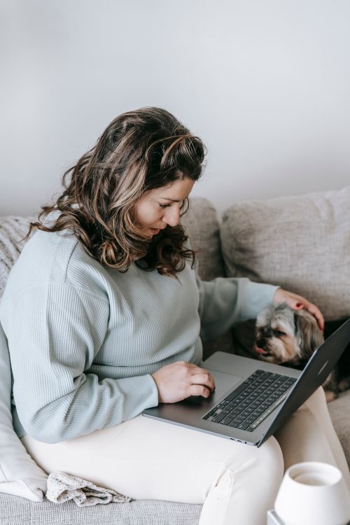 Free Woman sitting on a couch using a laptop, accompanied by a small dog, showcasing a comfortable work-from-home setting. Stock Photo