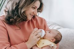 Optimistic crop mother with curly hair feeding infant baby with bottle while sitting on comfortable couch in light living room at home