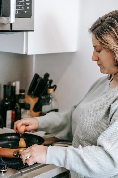 Side view of crop focused female breaking egg into frying pan while cooking breakfast on stove in light kitchen at home