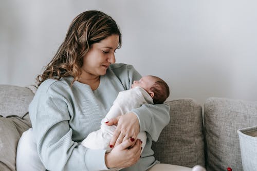 Free Side view of caring mother with infant baby in arms sitting on comfortable couch in light living room at home Stock Photo