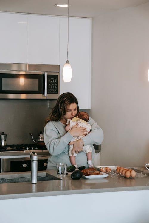 Free Loving young mother with long hair in casual clothe hugging cute baby while standing in kitchen during breakfast preparation Stock Photo