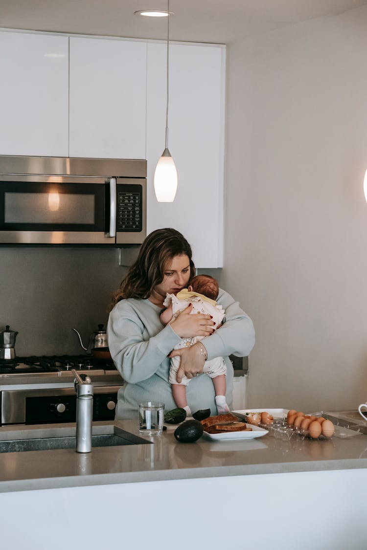 Young Mom Embracing Baby And Cooking In Kitchen