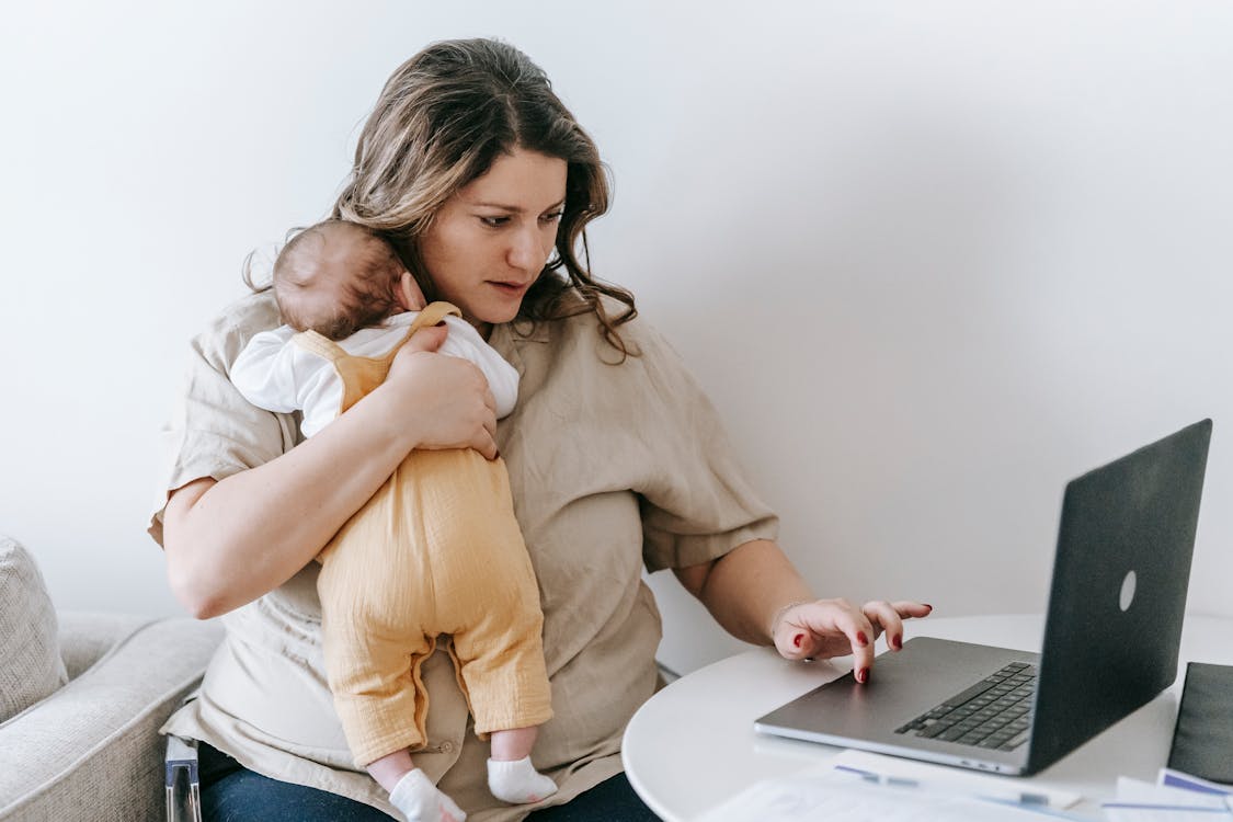 Concentrated young female freelancer embracing newborn while sitting at table and working remotely on laptop at home