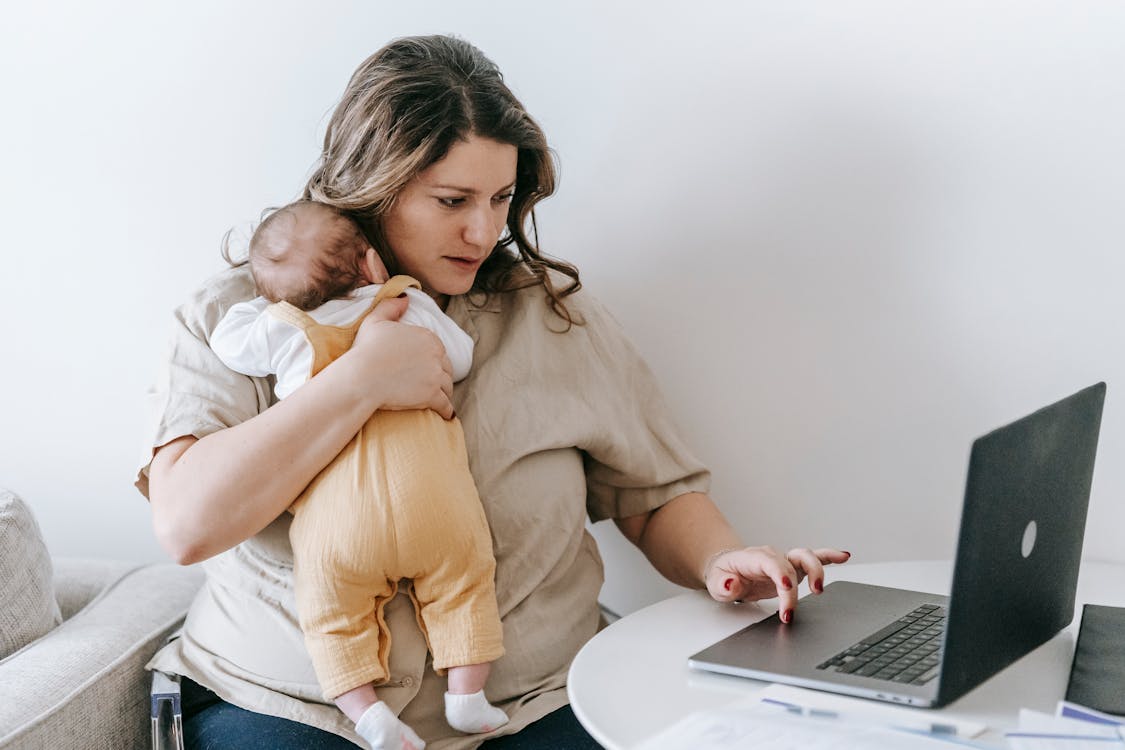 Subway Concentrated young female freelancer embracing newborn while sitting at table and working remotely on laptop at home